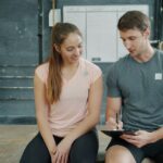 Profissional de Educação Física conversando com aluna e registrando informações de avaliação em uma prancheta